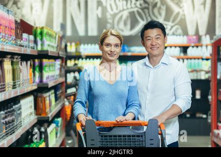 Ritratto di acquirenti felici in supermercato, coppia interrazziale asiatico uomo e donna, sorridente e guardando la macchina fotografica, tra scaffali con merci Foto Stock