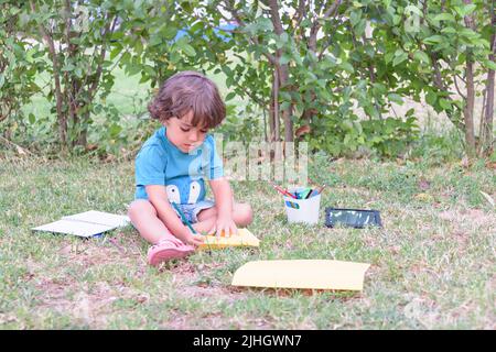 Il ragazzino sta usando una penna magica per disegnare immagini in un libro su un tavolo di legno nel parco con espressione facciale di determinazione. Foto Stock