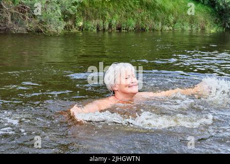 Donna anziana felice con capelli grigi godendo di nuoto in un fiume in campagna. Foto Stock