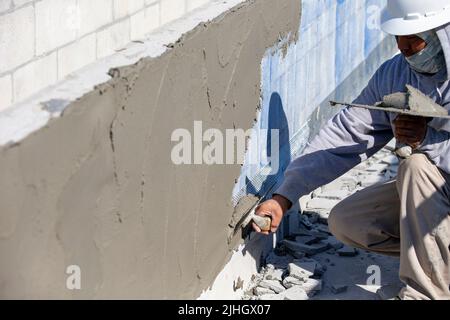 Un operaio di costruzione sta applicando fango di stucco su un blocco di cinderblock - parete del blocco di calcestruzzo, preparato con una soluzione appiccicosa blu ad esso aderisce al muro. Foto Stock