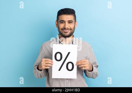 Soddisfatto uomo d'affari positivo tenendo carta e mostrando con segno percentuale iscrizione, guardando la macchina fotografica con sorriso, indossando camicia a righe. Studio interno girato isolato su sfondo blu. Foto Stock