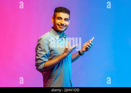 Vista laterale di un uomo ottimista felice in camicia utilizzando il telefono cellulare, sorridendo, mostrando il suo telefono cellulare, godendo l'app mobile. Studio interno girato isolato su colorato sfondo di luce al neon. Foto Stock