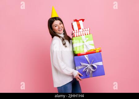 Sorridente bruna femmina positiva nel cono festa che celebra il compleanno, tenendo un sacco di scatole regalo, indossando un pullover bianco casual stile. Studio interno girato isolato su sfondo rosa. Foto Stock