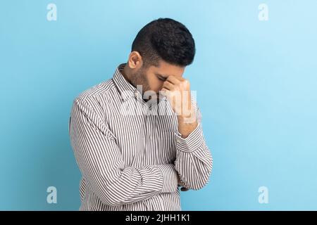 Depresso disperato uomo d'affari che sfrega gli occhi stanchi, piangendo a causa di disperazione e solitudine, rottura nervosa, indossare camicia a righe. Studio interno girato isolato su sfondo blu. Foto Stock