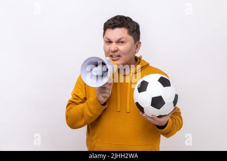 Ritratto di uomo di mezza età che tiene calcio palla bianca e nera e megafono, facendo annuncio sulla partita di calcio, indossando felpa con cappuccio urban style. Studio interno girato isolato su sfondo bianco. Foto Stock