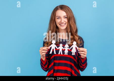 Gentile donna amichevole indossando a strisce stile casual maglione, tenendo in mano la catena di carta persone, concetto di famiglia felice, paternità, infanzia. Studio interno girato isolato su sfondo blu. Foto Stock