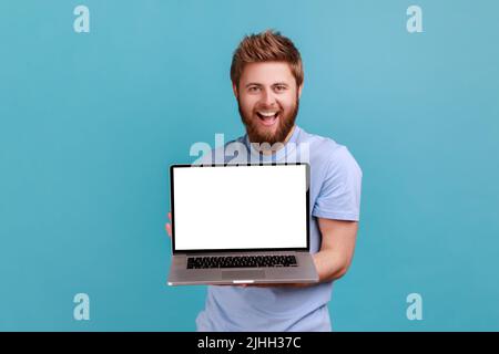 Ritratto di uomo barbato che tiene e mostra laptop con display vuoto vuoto, posto per la tua pubblicità, guardando la fotocamera, sorriso toothy. Studio interno girato isolato su sfondo blu. Foto Stock