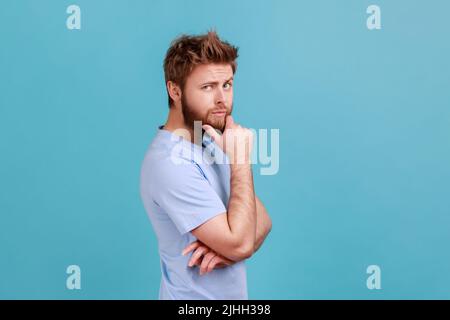 Ritratto di vista laterale del giovane uomo bearded premuroso che tocca il mento mentre si medita il piano, avendo dubbi circa scelta difficile, non sicuro. Studio interno girato isolato su sfondo blu. Foto Stock