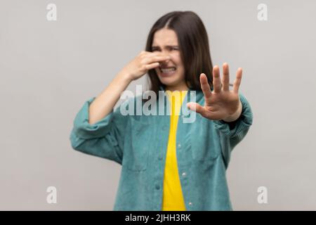 Odore terribile. Donna confuso pizzicando il naso per trattenere il respiro e mostrando il gesto di arresto, travestito da odore sgradevole, freccette, indossando giacca casual stile. Studio interno girato isolato su sfondo grigio. Foto Stock