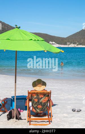 Uomo con cappello da sole su lettini colorati accanto all'ombrello, sulle spiagge paradisiache di Pontal do Atalaia Cabo Frio, Brasile. Foto Stock