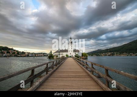 Schloss Ort sul lago Traunsee vicino a Gmunden, Austria. La vista sul molo di legno che conduce verso la chiesa. Cielo nuvoloso con tramonto sullo sfondo. Foto Stock