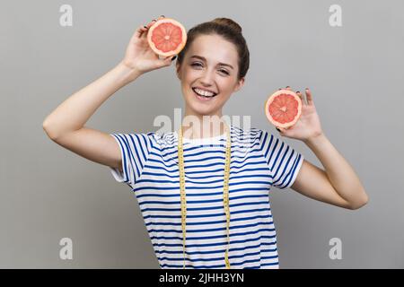 Ritratto di gioiosa felice donna ottimista indossando una T-shirt a righe in piedi con fette di pompelmo e nastro di misurazione, sorridendo alla fotocamera. Con studio interno girato isolato su sfondo grigio. Foto Stock