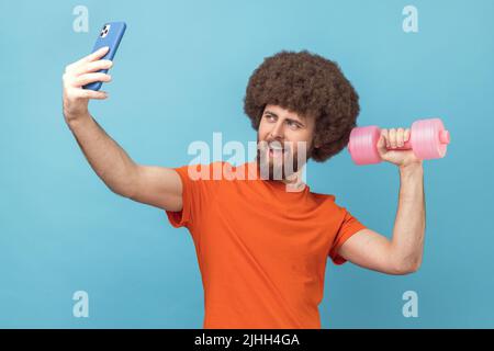 Uomo positivo con acconciatura Afro con T-shirt arancione per videochiamare e braccio sollevato con mubell, per prendere selfie o trasmettere in live streaming. Studio interno girato isolato su sfondo blu. Foto Stock