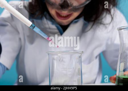 Primo piano di un operatore di laboratorio che gocciola gocce di sangue in un bicchiere di vetro durante l'esecuzione di test pericolosi. Biochimico maniacale dall'aspetto sciocco lunatico che utilizza il contagocce per sperimentare composti chimici tossici. Foto Stock