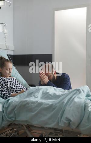 Il padre preoccupato prega per la figlia ammalata che riposa sul letto del paziente. Ammalato bambina sotto trattamento medico che riposa all'interno del reparto pediatria ospedale mentre il genitore prega per la sua salute. Foto Stock