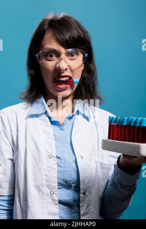 Donna biochimica con tubo di vetro tra i denti mentre tiene il vassoio da laboratorio con sostanza liquida rossa. Specialista di microbiologia con vetreria da laboratorio con provette riempite con campioni di sangue. Foto Stock