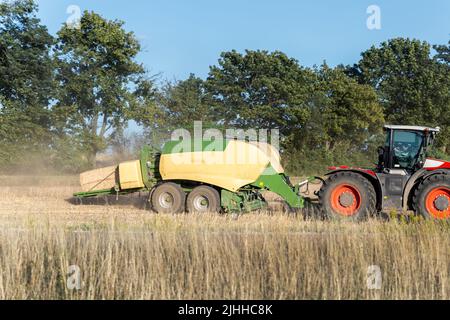 Vista laterale della macchina motrice moderna di grandi dimensioni con rimorchio per imballatrice che blocca il fieno dopo la mietitura su campo del grano. Attrezzatura agricola e agribuisness Foto Stock