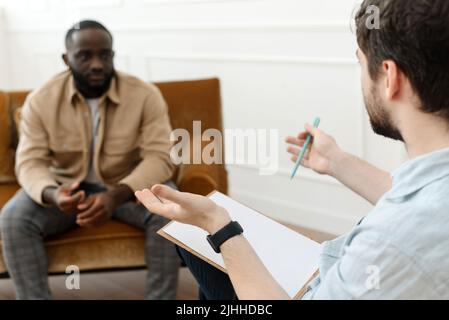 Il terapeuta prende appunti mentre parla con un paziente depresso di sesso maschile e parla con un paziente afroamericano. Disperato uomo nero che soffre di nervi Foto Stock