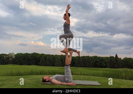 Giovane coppia che fa acro yoga nel parco. Uomo sdraiato sull'erba e donna equilibrante nei suoi piedi Foto Stock