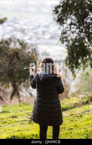 Violinista femminile di bruna visto dalla parte posteriore che suona il violino fuori nella montagna. Verticale Foto Stock