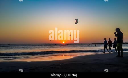 Kite surf al tramonto. Kite boarding attività sportive all'aperto. La gente silhouette camminare sulla spiaggia sabbiosa all'alba con il sole arancione e il cielo colorato Foto Stock