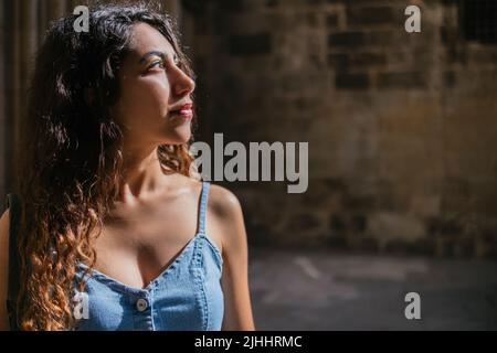 Giovane turista ispanico femmina con capelli ricci guardando via mentre in piedi alla luce del sole in camera oscura di vecchio edificio Foto Stock