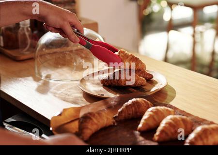 Immagine closeup del barista della caffetteria che mette i croissant caldi appena sfornati dal foglio di cottura al piatto da portata Foto Stock