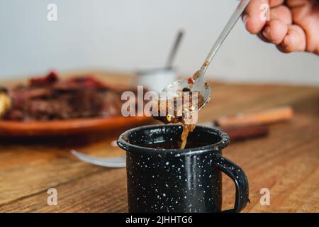 Dettaglio di una piccola bottiglia di salsa fatta in casa presa da un cucchiaio. Foto di alta qualità. Una mano che usa il cucchiaio per servire la salsa. Piatto di carne sullo sfondo. Tavola di legno sullo sfondo Foto Stock