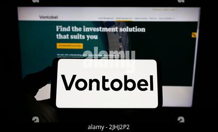 Persona che tiene il cellulare con il logo della società finanziaria svizzera Vontobel Holding AG sullo schermo di fronte al sito web aziendale. Mettere a fuoco sul display del telefono. Foto Stock