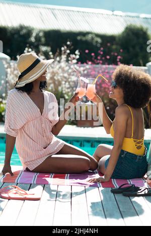 Le amiche biraciali tostano le bevande mentre si siedono su un asciugamano e si godono le feste in piscina in estate Foto Stock