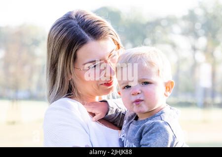 Felice madre giovane con un bambino piccolo o bambino nelle sue braccia, abbracciante e coccolante, in piedi nel mezzo di un parco ai raggi del sorgere del sole del mattino. Foto di alta qualità Foto Stock