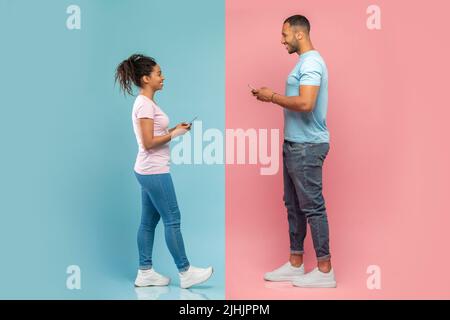 Coniugi afro-americani positivi che tengono smartphone e si guardano l'un l'altro, sfondo blu e rosa, vista laterale Foto Stock