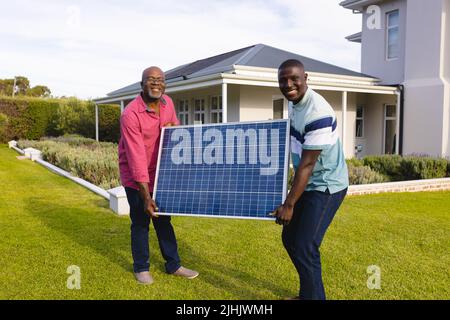 African american sorridente uomo anziano e figlio che trasportano il pannello solare mentre si alza sul campo in iarda Foto Stock