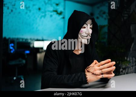 Uomo hacker pericoloso e irriconoscibile indossando maschera anonima e felpa con cappuccio che tiene i cordoni nelle mani e guardando via seduto al tavolo nella stanza del seminterrato Foto Stock