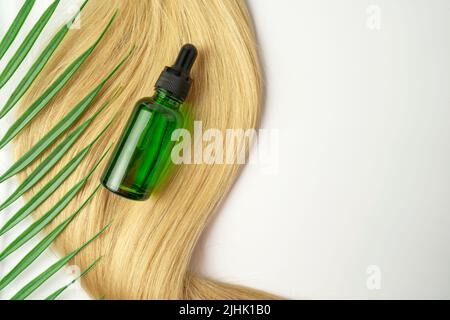 Un flacone contagocce di olio verde CBD o siero per la cura dei capelli in capsule dorate adagiate su una ciocca di capelli biondi. Cura dei capelli e concetto di lisciatura Foto Stock