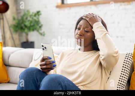 Delusa la giovane donna afroamericana che tiene lo smartphone e guarda lo schermo del telefono seduto sul divano a casa, ha ricevuto una cattiva notifica o messaggi, si sente turbata Foto Stock