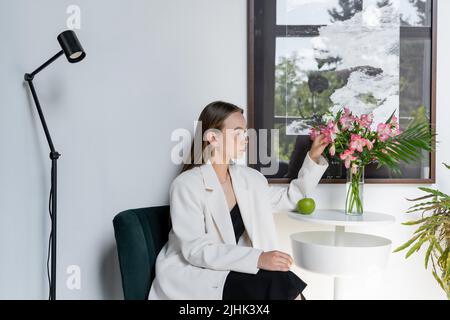 donna in blazer bianco che tocca i fiori di alstroemeria sul tavolino da caffè vicino alla finestra Foto Stock