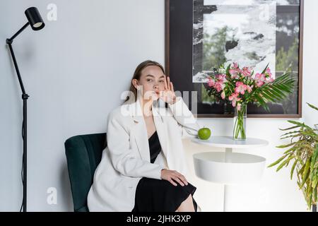 donna elegante in blazer bianco seduto vicino a fiori di mela e di alstroemeria su tavolino da caffè Foto Stock