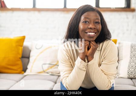 Bella donna afroamericana seduta sul comodo divano in accogliente soggiorno, donna multirazziale che riposa a casa, posa e guarda la macchina fotografica Foto Stock