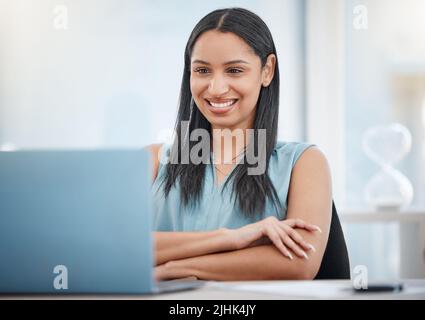 Felice giovane gara mista donna d'affari sorridente mentre si gode di lavorare su un computer portatile seduto su una sedia in un ufficio al lavoro. Un boss ispanico femmina Foto Stock