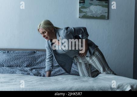 donna bionda che entra a letto mentre sente dolore allo stomaco durante la menopausa Foto Stock