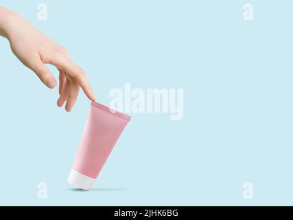 La mano di Womans tocca un tubo rosa di crema cosmetica su sfondo blu chiaro Foto Stock