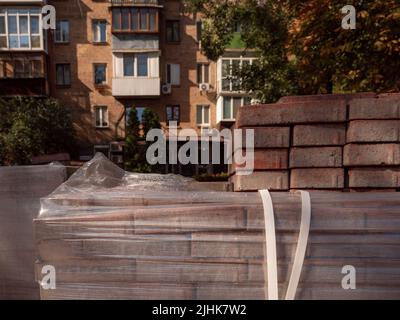 Una pila di mattonelle rosse in primo piano e un vecchio edificio di mattoni rossi illuminato dal sole sullo sfondo a Kyiv, Ucraina. Strada in costruzione Foto Stock