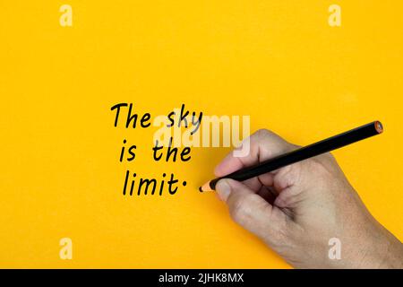 La mano maschile scrive a matita nera la parola IL CIELO È IL LIMITE su uno sfondo giallo. Parole di incoraggiamento e concetto di business. Foto Stock