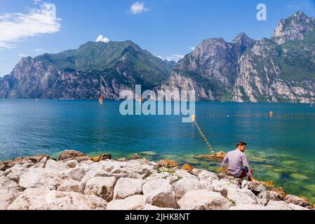 Uomo seduto accanto al Lago di Garda mentre i windsurf navigano sul lago. Nago-Torbole è un comune del Trentino, in Trentino Foto Stock