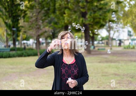 Donna trans matura che soffia bolle in un parco Foto Stock