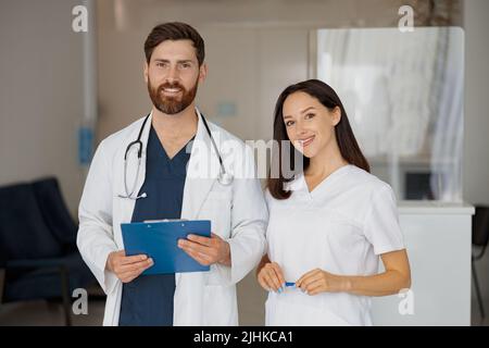 Ritratto di felice medico e infermiere in piedi vicino alla reception in ospedale e guardando la macchina fotografica Foto Stock