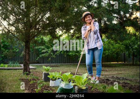 Affascinante donna afroamericana, il giardiniere sorride alla macchina fotografica, in posa con una zappa mentre lavora nell'orto Foto Stock