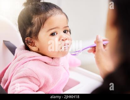 Una bambina di razza mista che viene nutrita con un cucchiaio da mamma a casa. Cute toddler con la sua bocca ripiena di porridge seduta in una sedia di alimentazione per godere Foto Stock