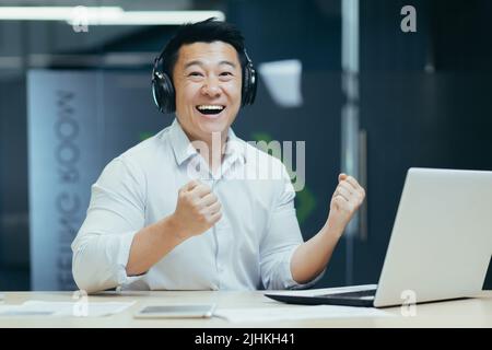 ritratto di uomo d'affari con cuffie, uomo asiatico sorridendo e guardando la macchina fotografica, boss guardare lo sport partita sul posto di lavoro Foto Stock